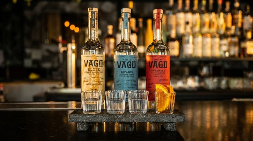 Three Vago Mezcal bottles and glasses with drinks and orange slices on a bar.