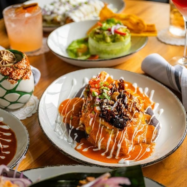 A wooden table laden with various colorful Mexican dishes and cocktails, including enchiladas and guacamole.