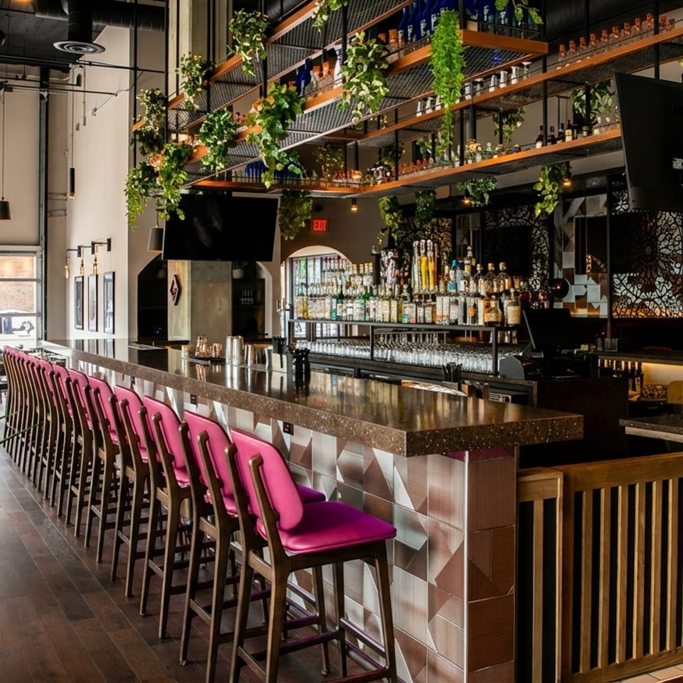 Modern bar area with pink stools, a fully stocked bar, and hanging plants, next to a dining space.