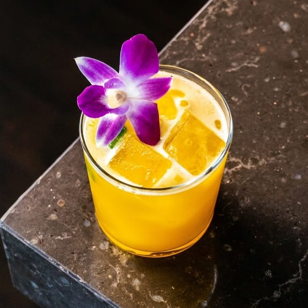 Vibrant yellow cocktail with ice cubes and a purple orchid garnish on a dark marble surface.