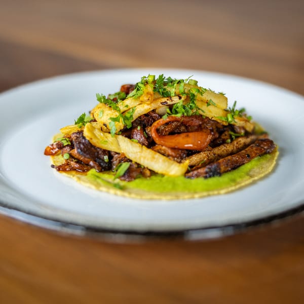 A single taco on a white plate, filled with meat, fries, and green sauce, garnished with parsley.