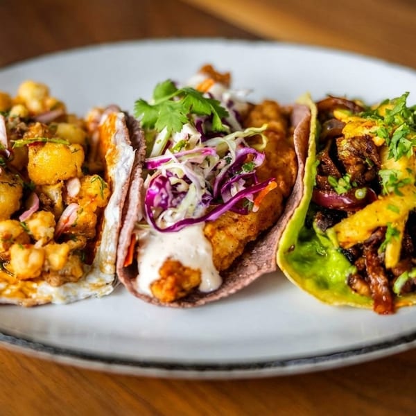 Three vibrant tacos with diverse fillings, including cauliflower, fish, and meat, on a white plate.