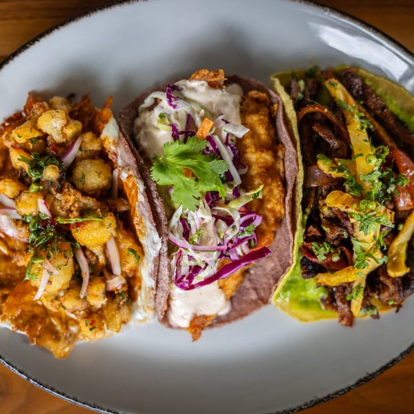 Three gourmet tacos with diverse fillings, including fried cauliflower, fish, and beef, on a white plate.