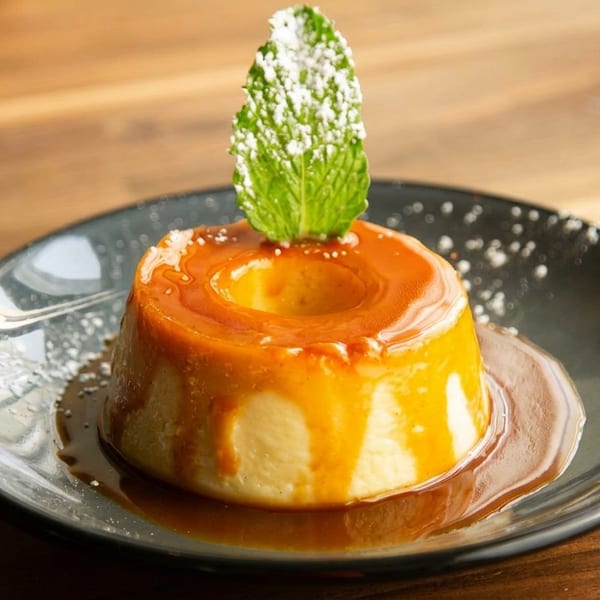 Flan dessert with caramel sauce, powdered sugar, and a mint leaf on a dark plate.
