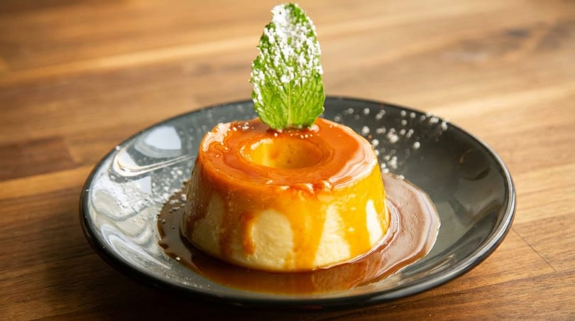 Flan dessert with caramel sauce, powdered sugar, and a mint leaf on a dark plate.
