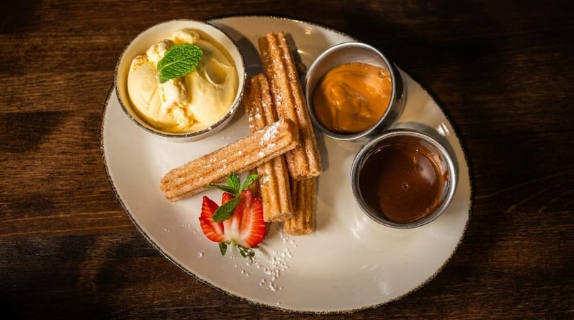 Churros with powdered sugar, vanilla ice cream, caramel, chocolate sauce, and strawberries on a plate.