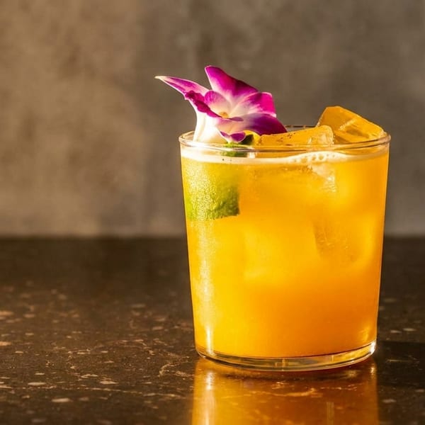 A vibrant orange cocktail with ice, garnished with a purple orchid and lime, on a dark reflective surface.