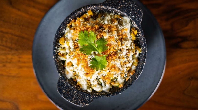 Elote, Mexican street corn, in a black bowl with cilantro, cheese, and breadcrumbs.