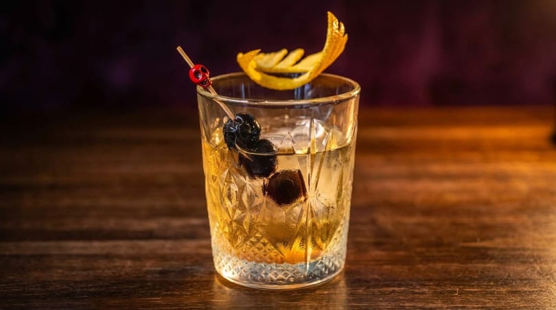 Old Fashioned cocktail with a skull pick, cherries, and orange peel on a wooden table.
